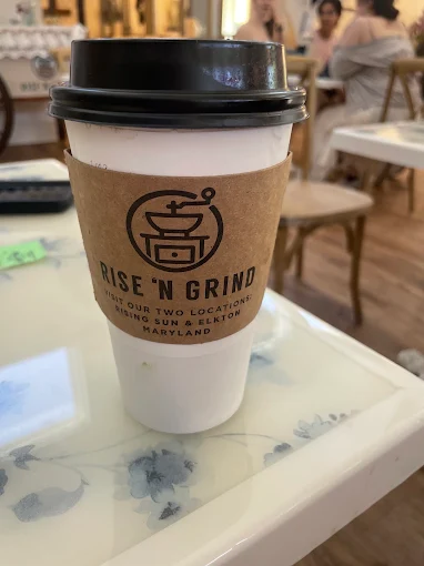 A to-go cup with a kraft sleeve printed with the Rise 'N Grind logo, sitting on a blue and white marbled table